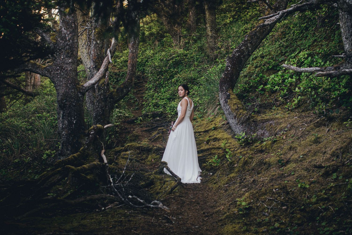 A figure in a flowing white dress stands in a mystical forest setting surrounded by mossy trees and natural foliage.