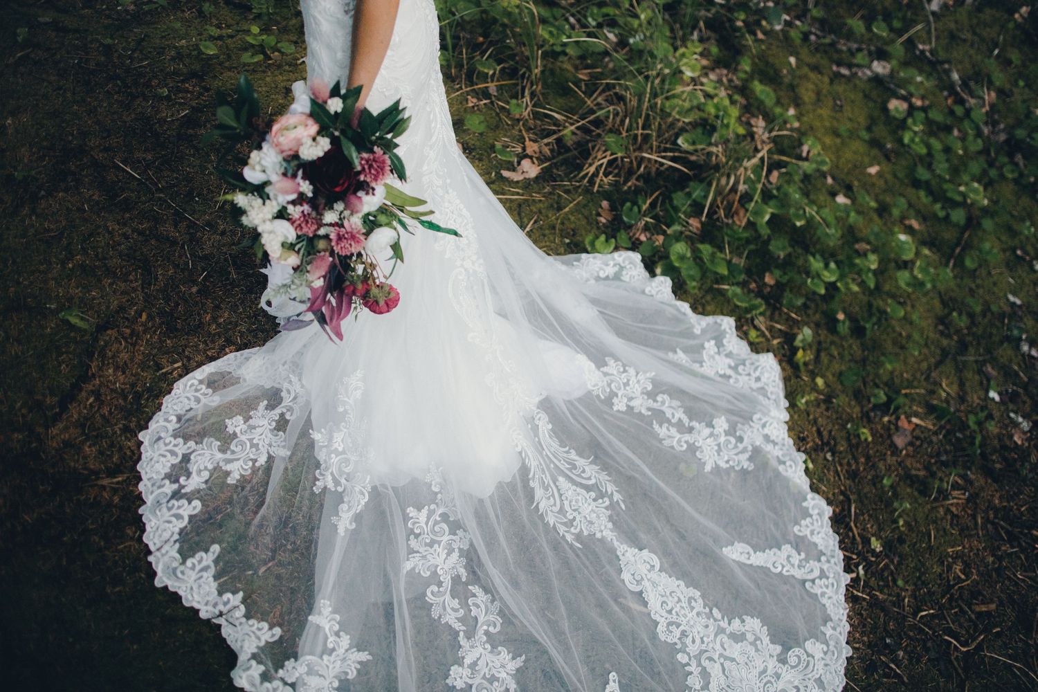 A flowing white lace wedding dress with delicate details spreads across grass while holding a pink and white bouquet.