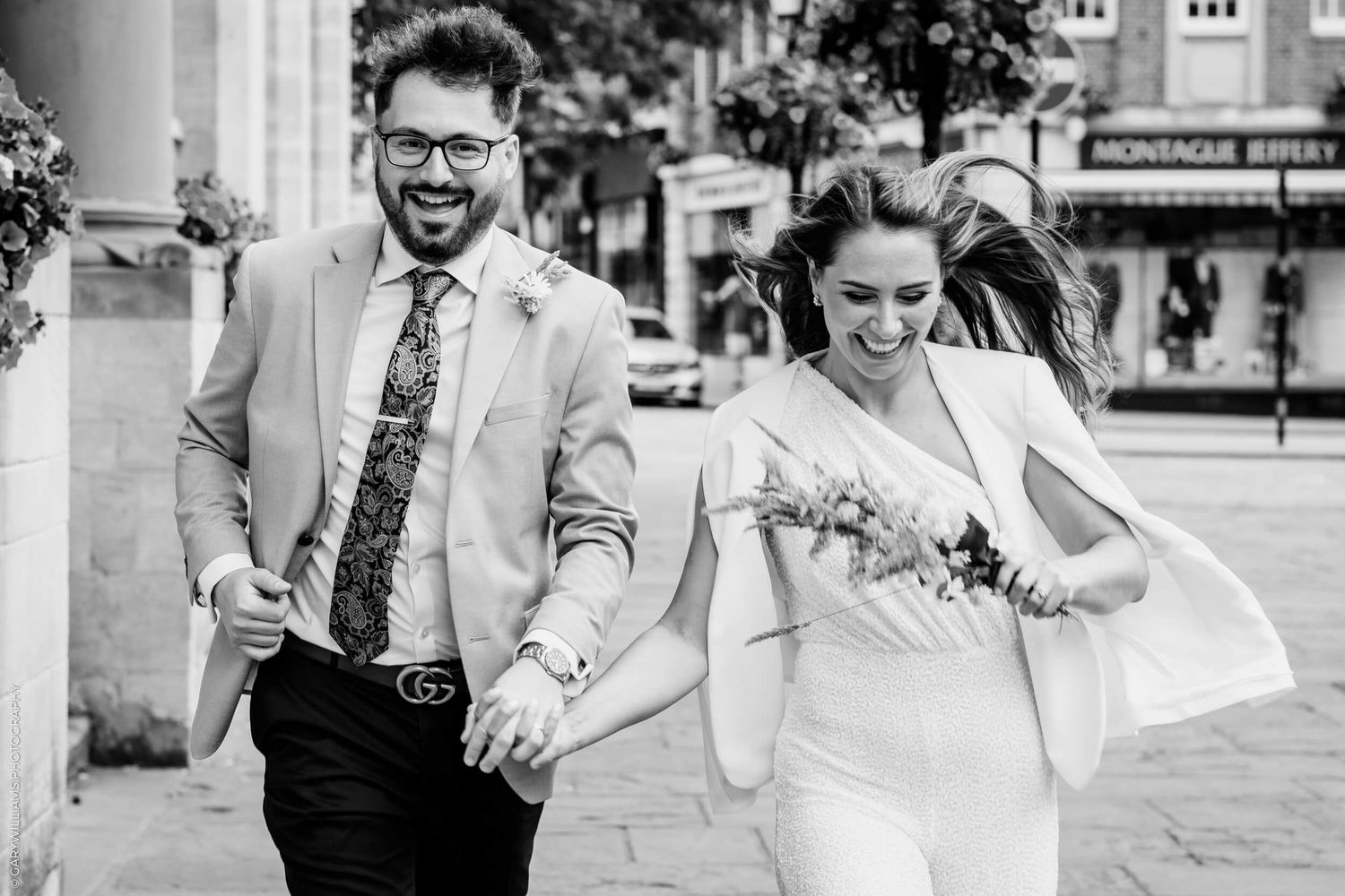 A happy couple walks hand in hand on their wedding day in black and white, with the bride in white and groom in a light suit.