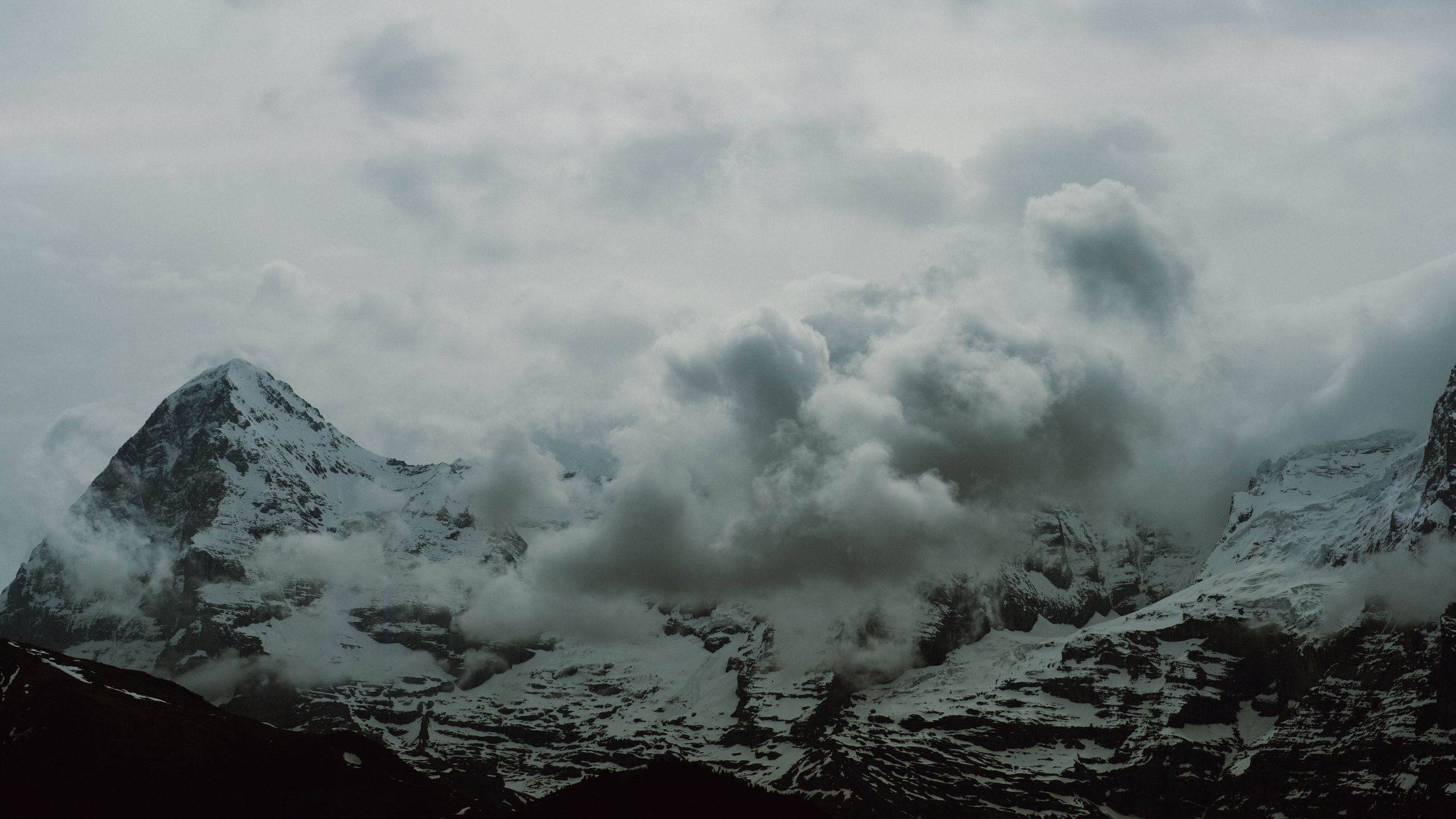 Snow-capped mountain peaks emerge through dramatic dark clouds in a moody alpine landscape.