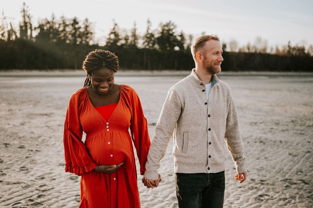 Maternity portrait of couple holding hands along kye bay beach in red dress during sunset Comox Valley Vancouver Island photographer