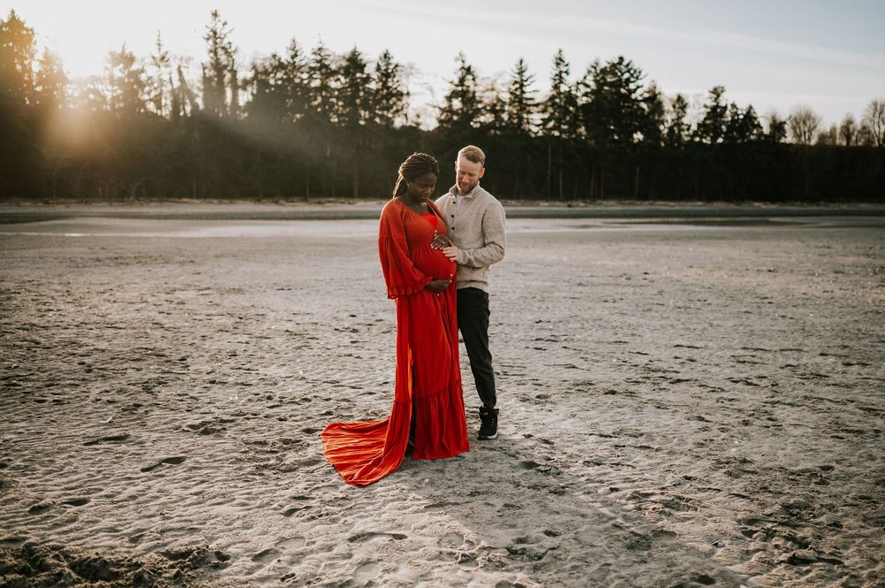 Maternity portrait of couple holding bump along kye bay beach in red dress by ocean during sunset Comox Valley Vancouver Island photographer