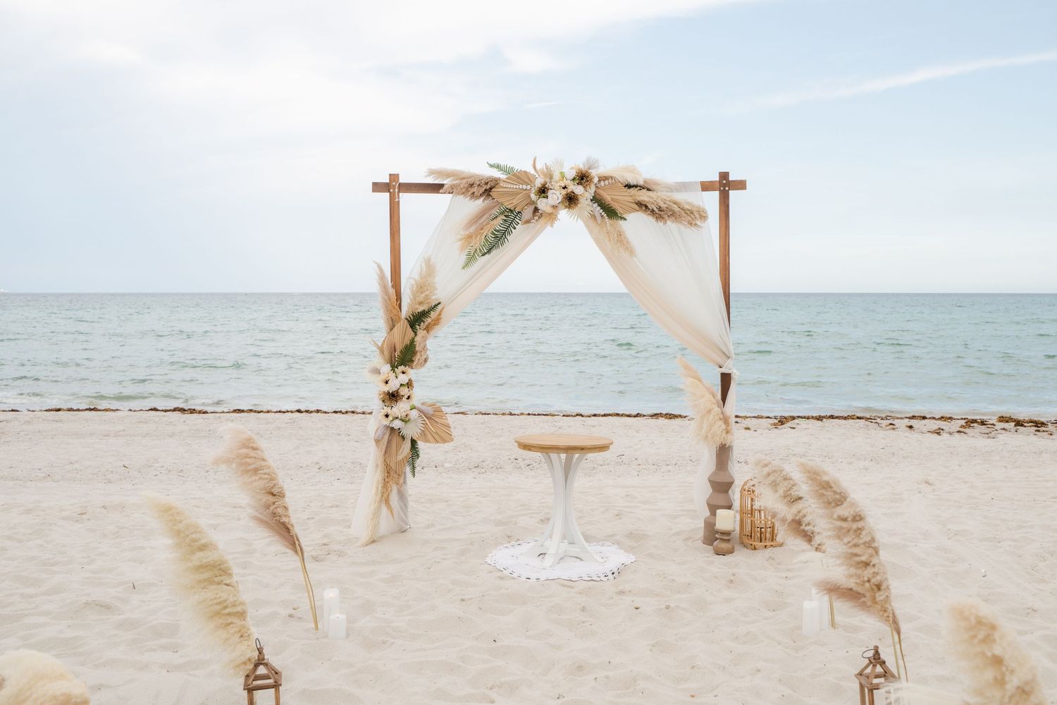 A romantic beach wedding ceremony setup with bamboo arch, white curtains, and boho dried palm decorations on sandy shores.