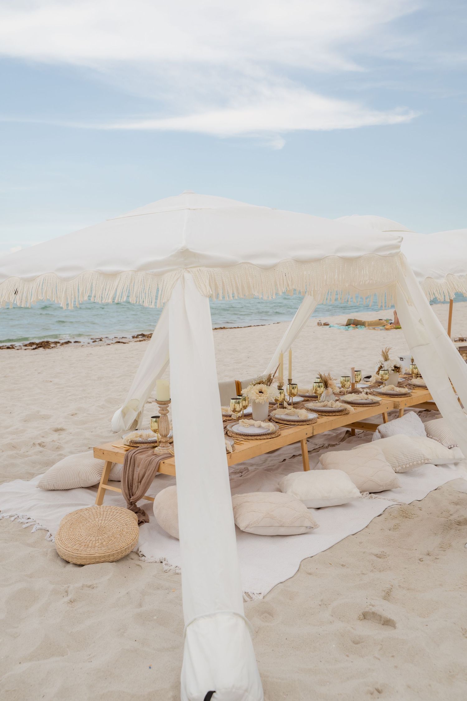 Elegant beach dining setup with white canopy and pillows on wooden table featuring coastal decor and place settings.