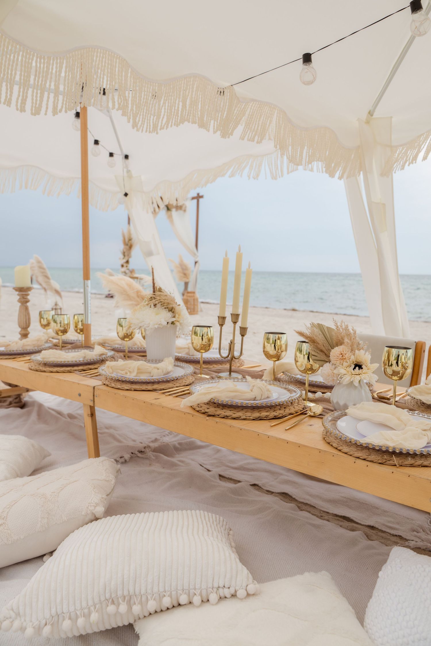 Elegant beachside dining setup with white fringe decor, taper candles, and floor cushions under a boho-style canopy.