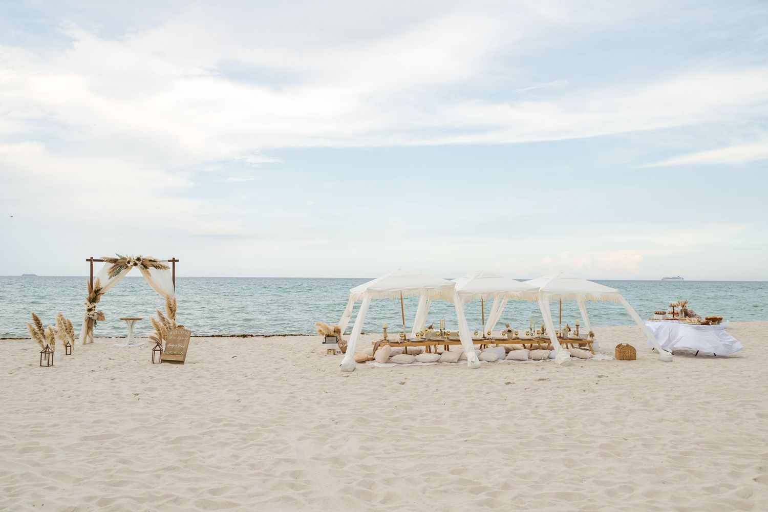 Romantic beach setup with white fabric canopies and seating arrangements along the shoreline on a sunny day.