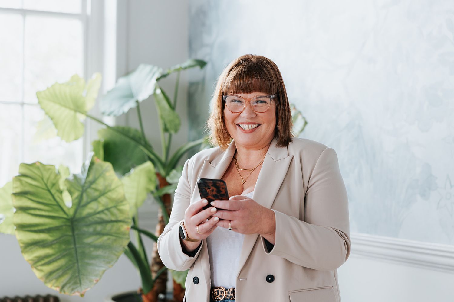 A happy woman in a beige blazer smiles while using her mobile phone next to large green indoor plants.