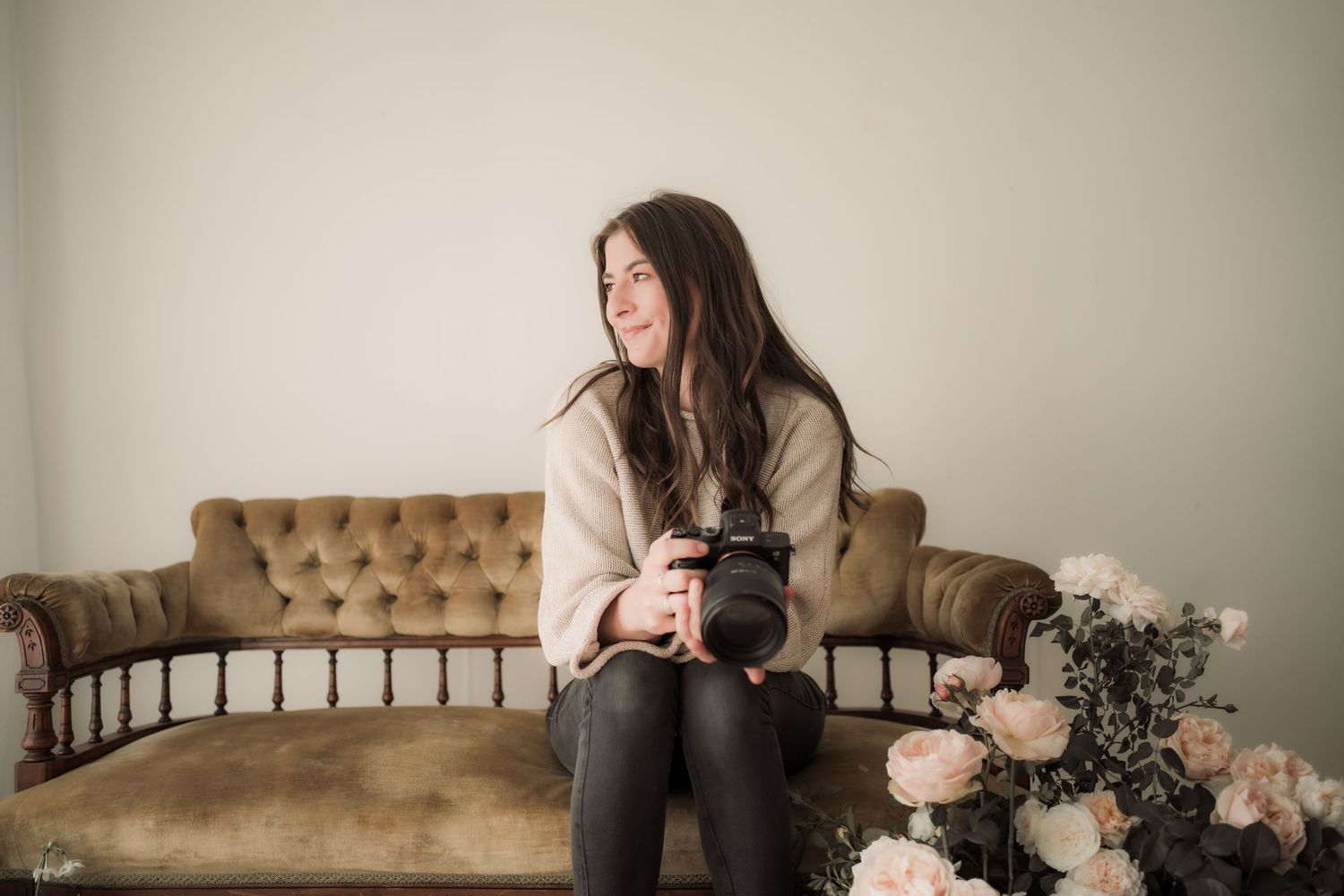 A photographer sits with a camera on a vintage tufted sofa beside pink roses and greenery in a minimalist setting.