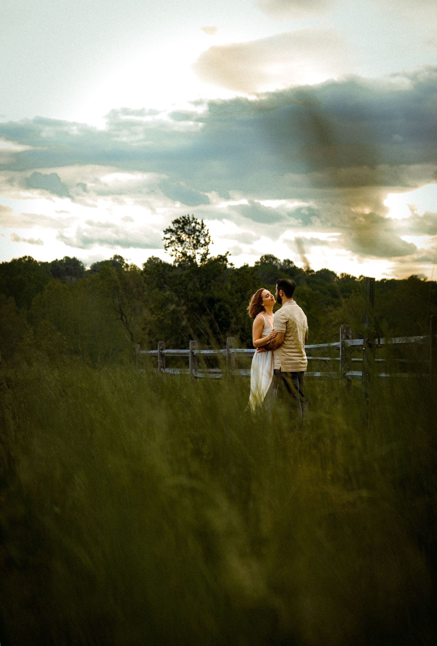 Two figures share a romantic moment in a meadow at sunset with a wooden fence and dramatic cloudy sky in the background.