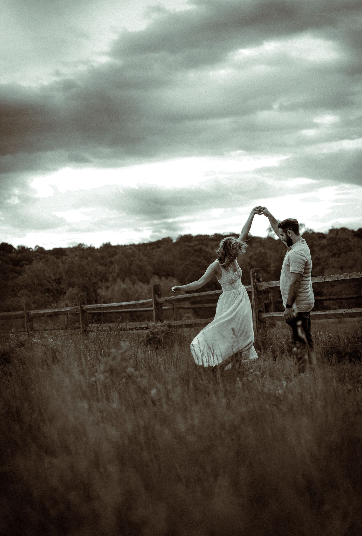 A couple dancing together in a meadow against a dramatic cloudy sky in black and white.