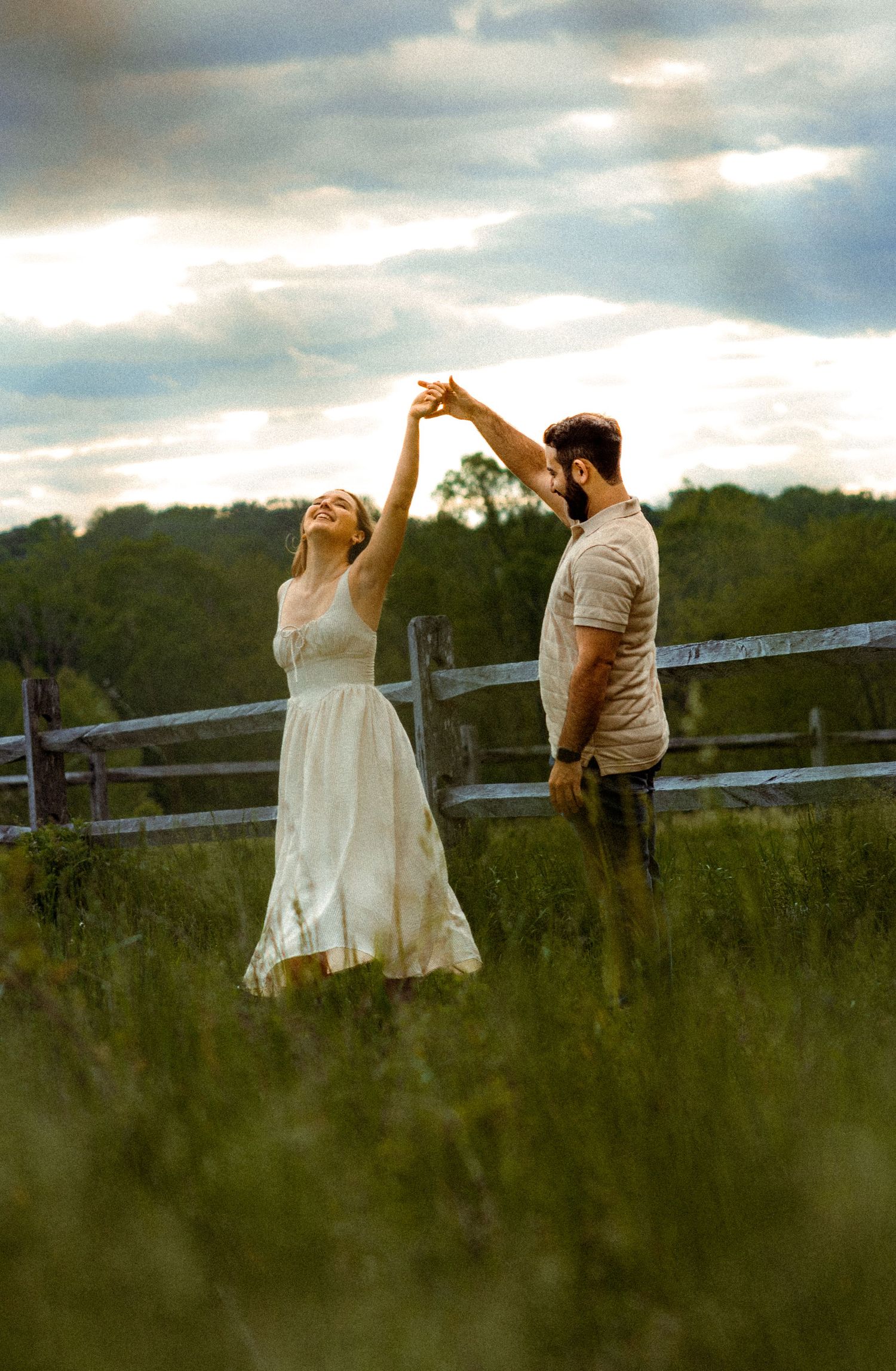 A couple dances together in a grassy meadow with a rustic wooden fence and dramatic cloudy sky behind them.