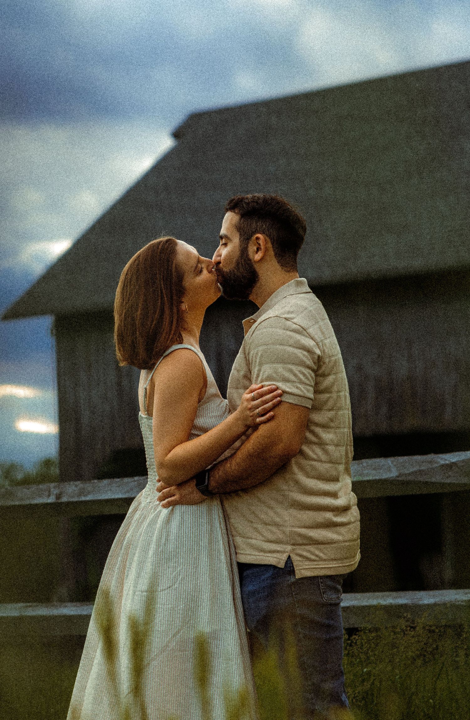 A romantic couple embraces by a rustic barn fence at dusk with dramatic cloudy skies in the background.