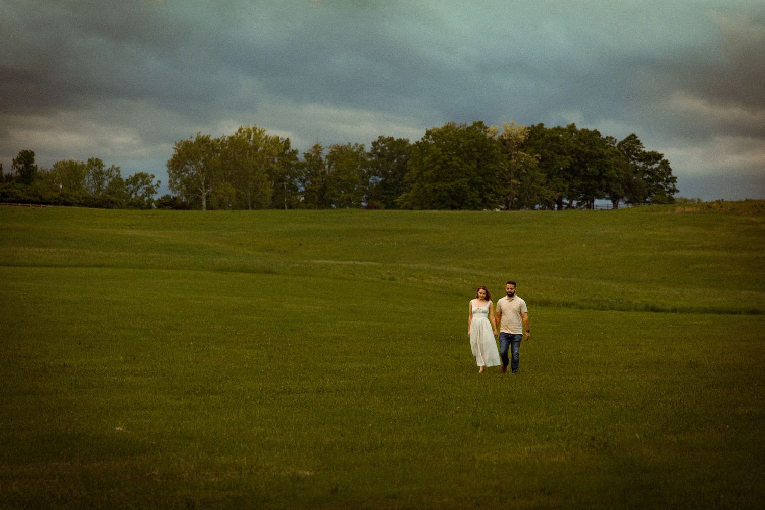 Two figures in light clothing walk together across a vast green field under moody storm clouds at dusk.