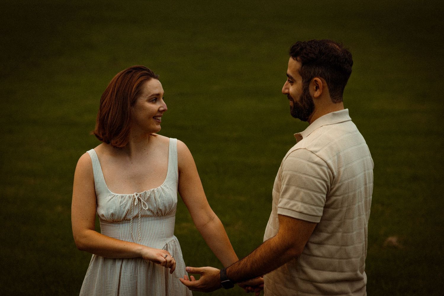 A couple laughs together while holding hands in a romantic outdoor setting with green grass at sunset.