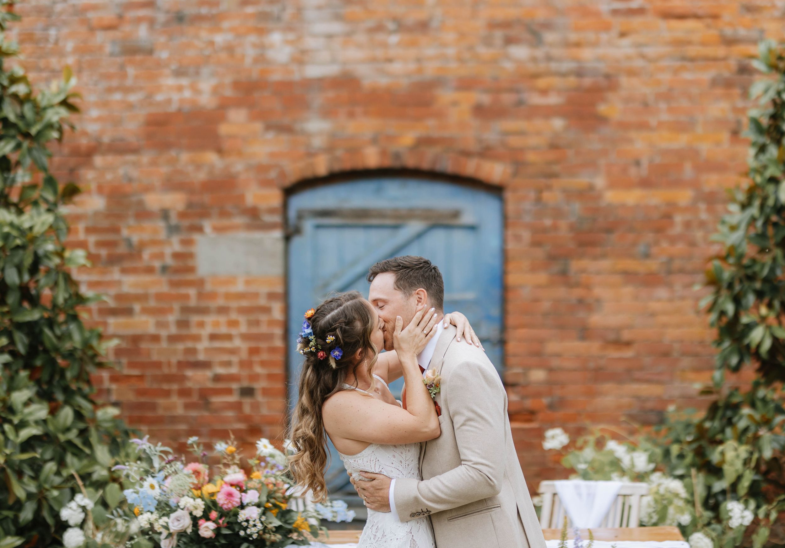 A romantic moment between a couple in wedding attire sharing a kiss against a rustic brick wall with climbing vines. Thorpe Garden.