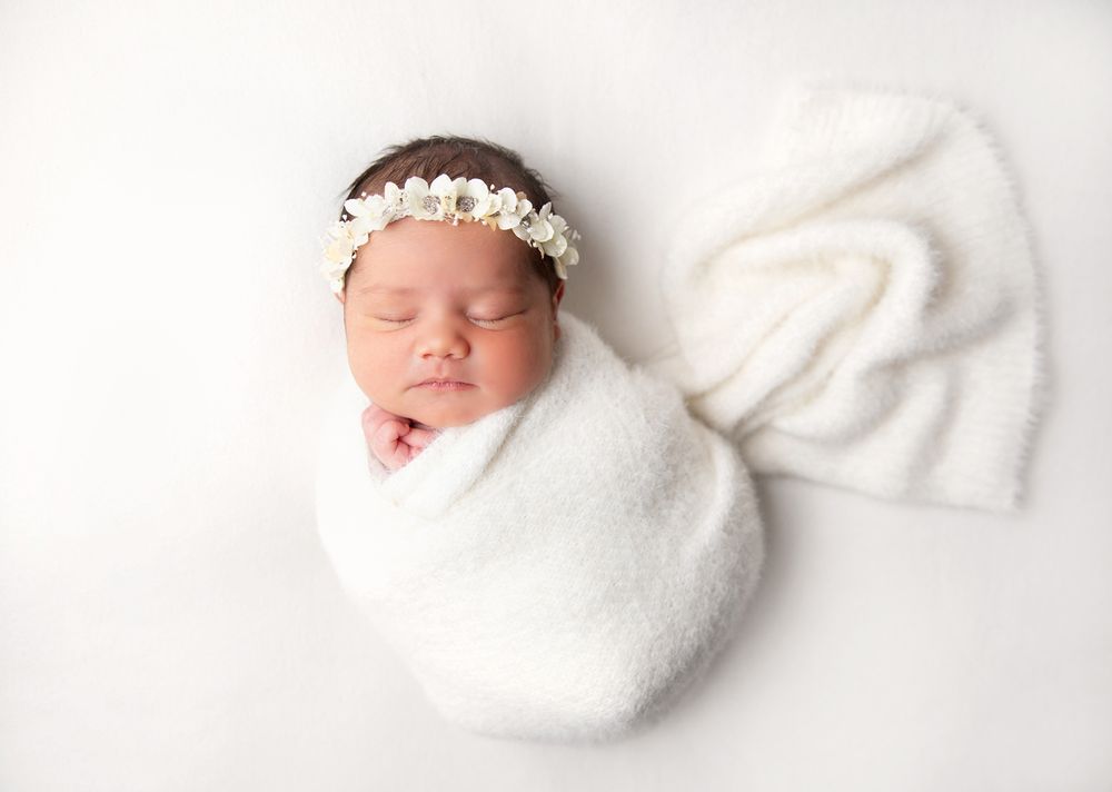 A sleeping newborn baby wrapped in a soft white blanket wearing a delicate floral headband. During a newborn session In London