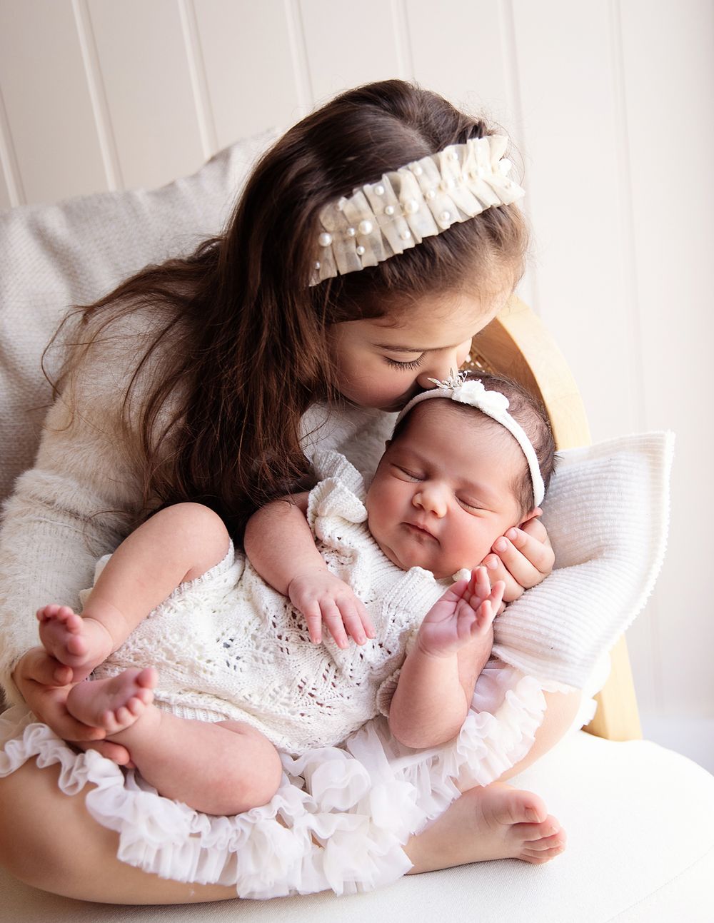 Siblings share a tender moment on white bedding, with both wearing matching white outfits and delicate headbands. During a newborn session
