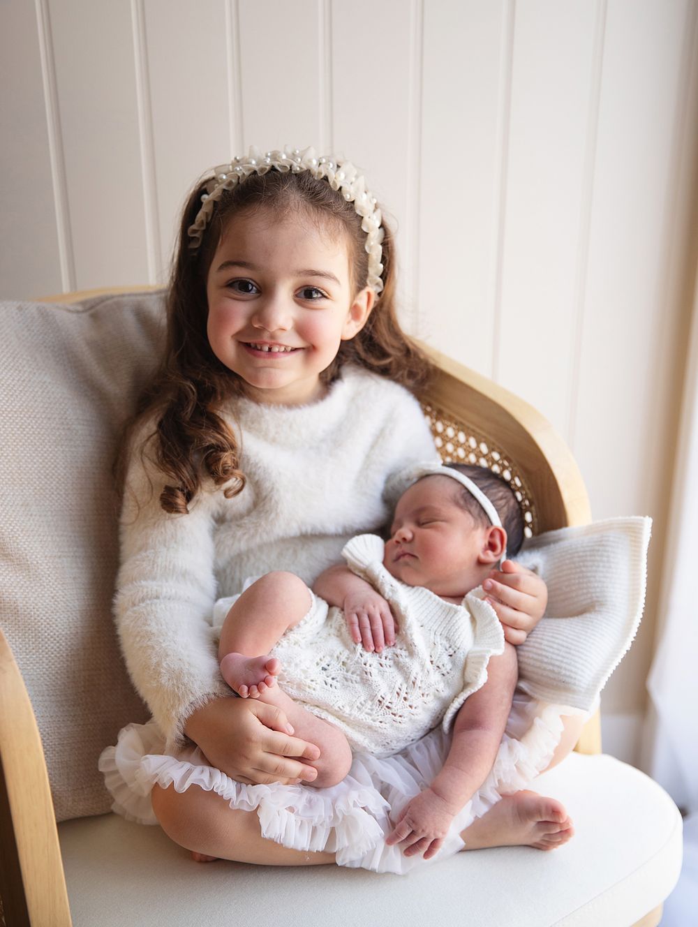 Two children sitting in a white cushioned chair, with the older sibling in a cozy sweater holding a newborn wearing a lace dress.