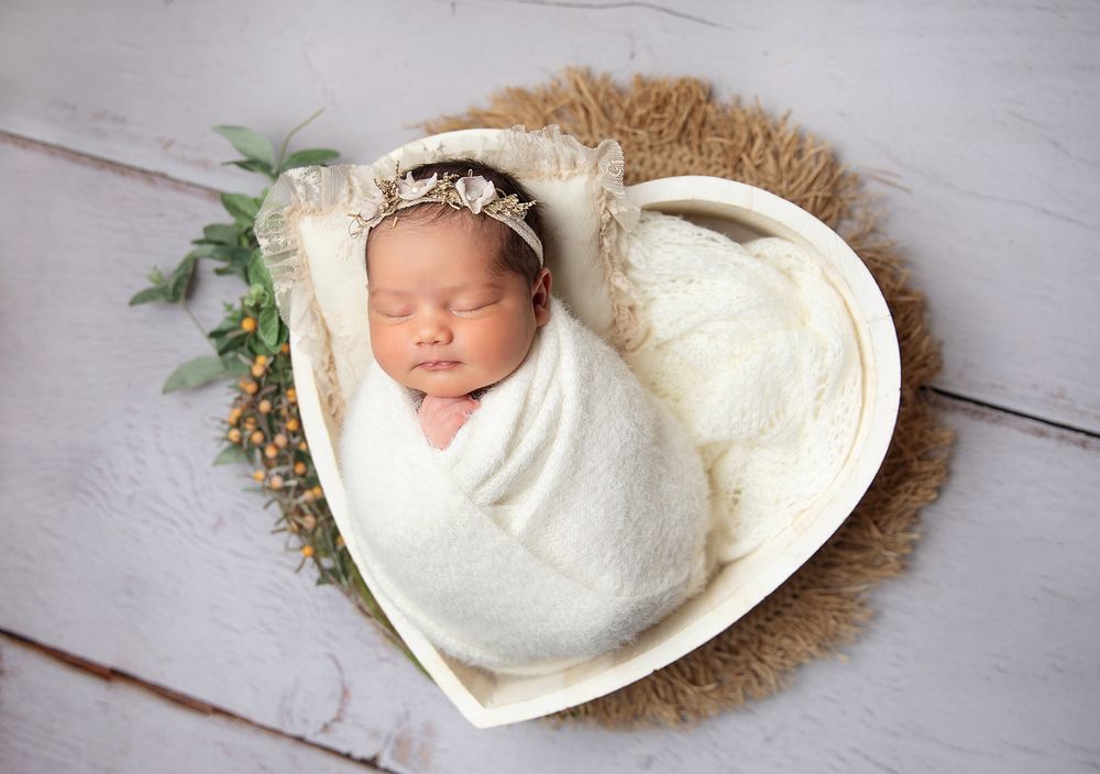 Sleeping newborn wrapped in white swaddle lying in heart-shaped basket with fur lining and greenery. During a newborn session In London