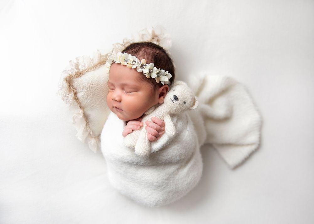 A sleeping newborn wrapped in white blanket wearing floral headband and holding a small bunny toy. During a newborn session In London