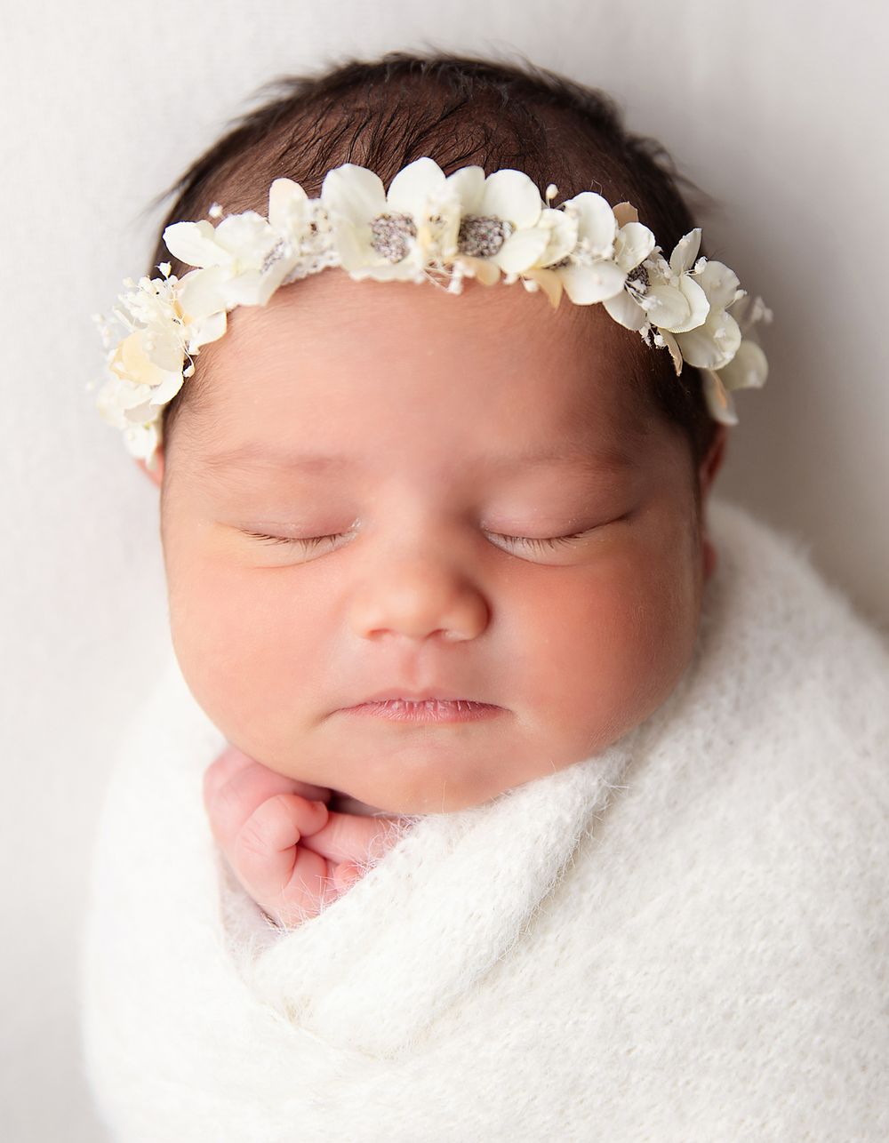 Sleeping newborn wrapped in white blanket wearing delicate floral headband with white and blush blooms in studio setting.
