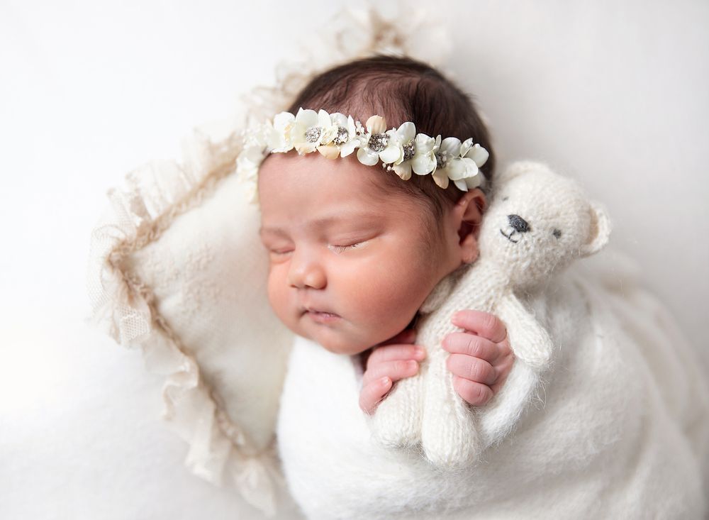 Peaceful sleeping newborn snugly wrapped in white with delicate floral headband holding small teddy bear. During a newborn session In London
