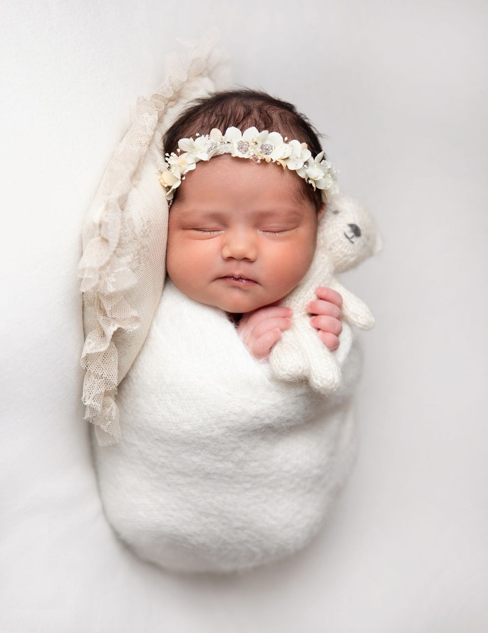 Sleeping newborn wrapped in soft white fabric wearing delicate floral headband and holding stuffed bunny. During a newborn session In London