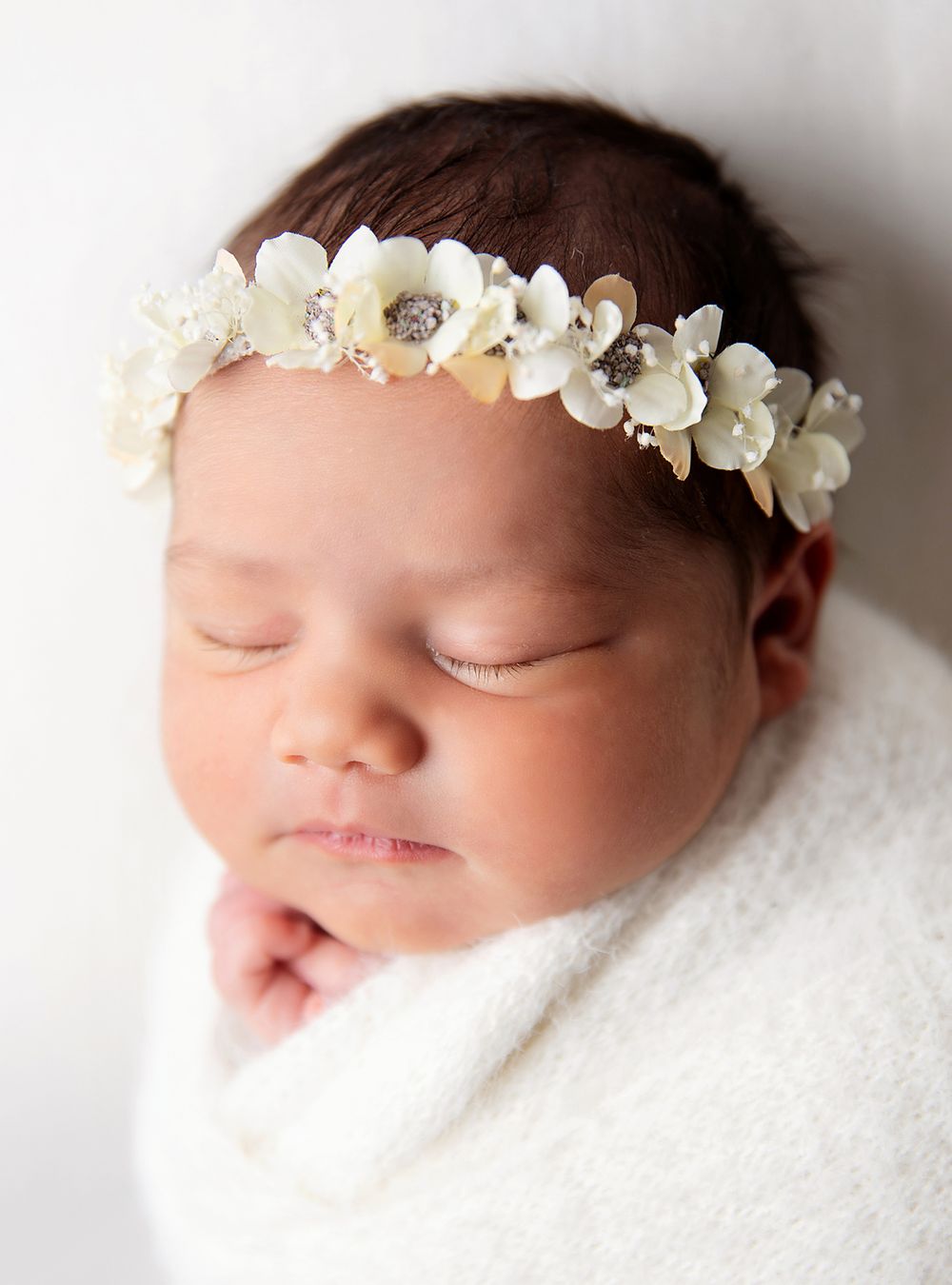Sleeping newborn wearing a white floral headband wrapped in soft white fabric on neutral background. During a newborn session In London
