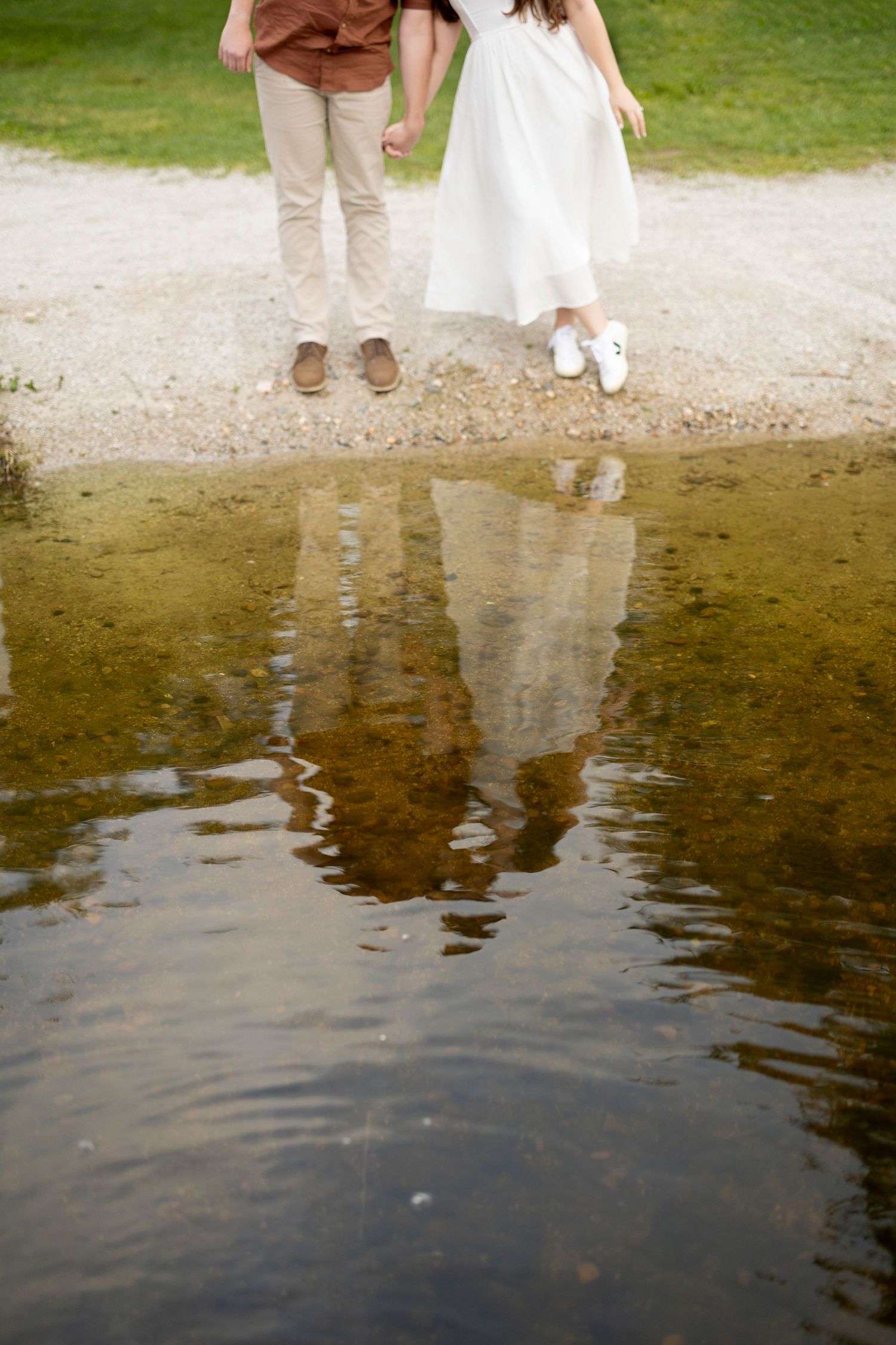 Reflection in still water shows partial view of two pairs of feet standing at waters edge in romantic pose.