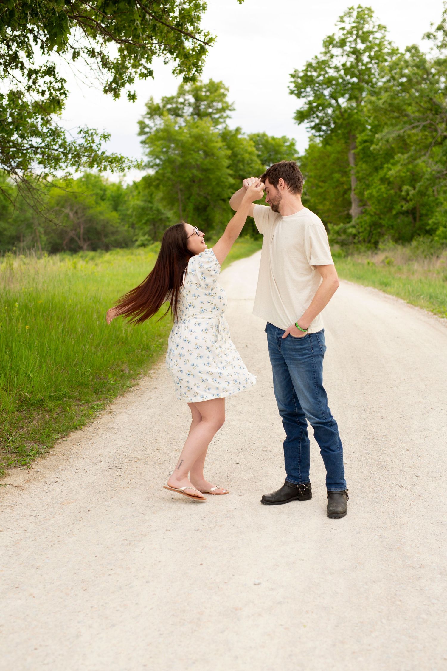 A couple shares a playful moment while dancing on a dirt path surrounded by green trees and grass.