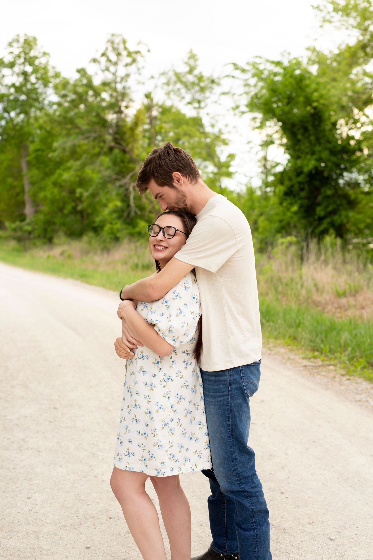 Couple embracing and smiling together on a dirt path surrounded by green trees and vegetation on a sunny day.