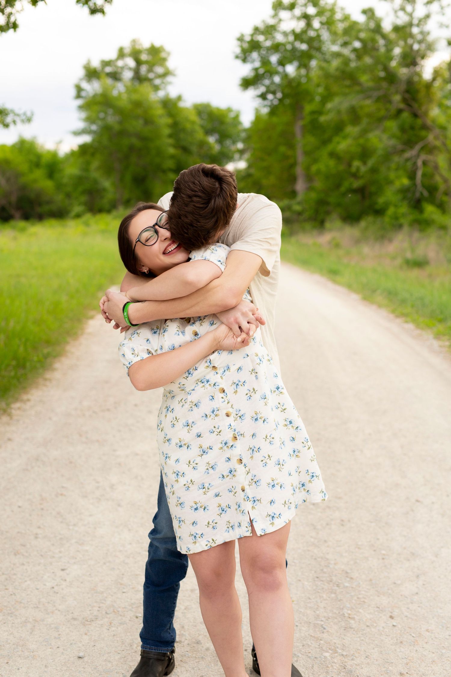 Two people share an embrace on a sunlit dirt path surrounded by green grass and trees.