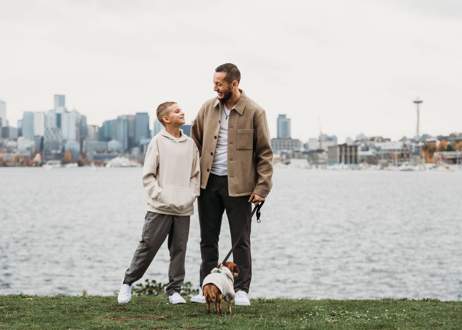 Two people stand with a small dog on grass by the waterfront with city skyline in background.