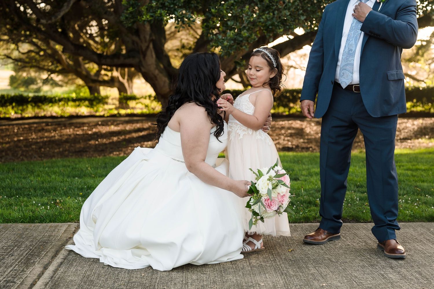 A sweet moment shared between a bride in a white gown and a small child in a matching dress in an outdoor park setting.