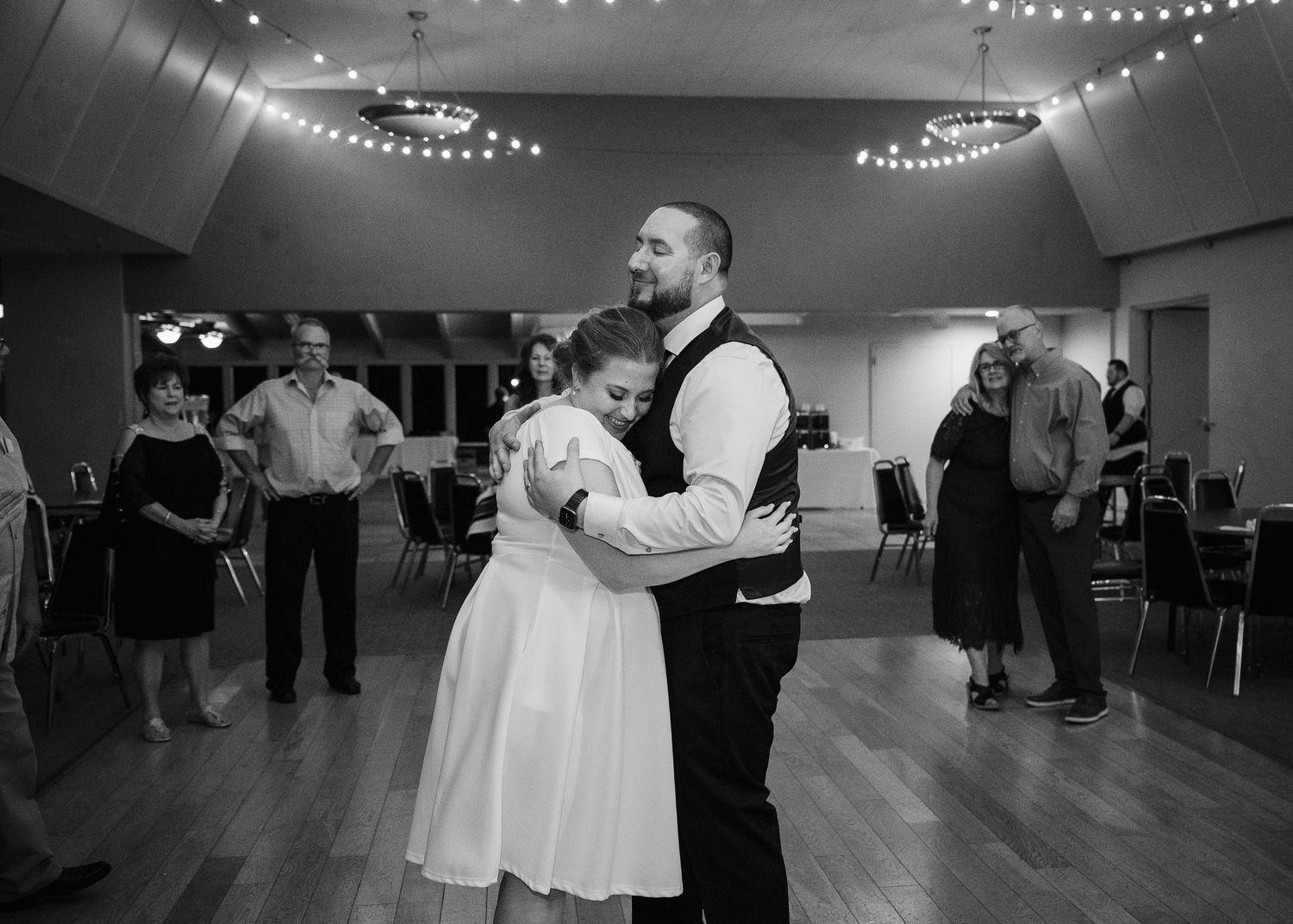 A couple dances together in a ballroom decorated with twinkling string lights during a romantic black and white moment.