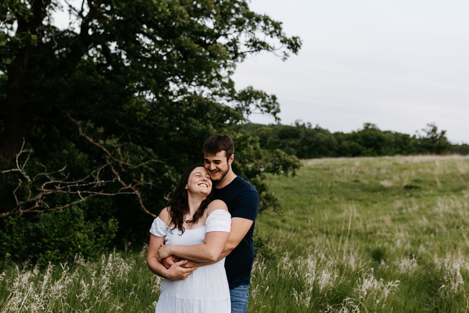 A couple embraces in a romantic moment while standing in a grassy meadow surrounded by trees on an overcast day.