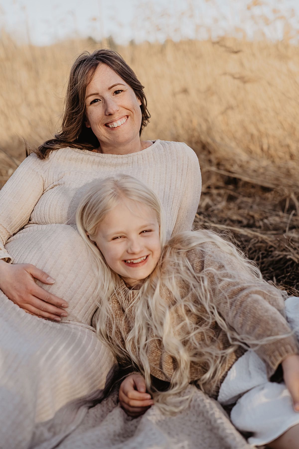 Gravid mamma ligger ner på en filt och hennes dotter lutar sig mot hennes mage, de ler och skrattar rakt in i kameran, fotograf Lets Bloom