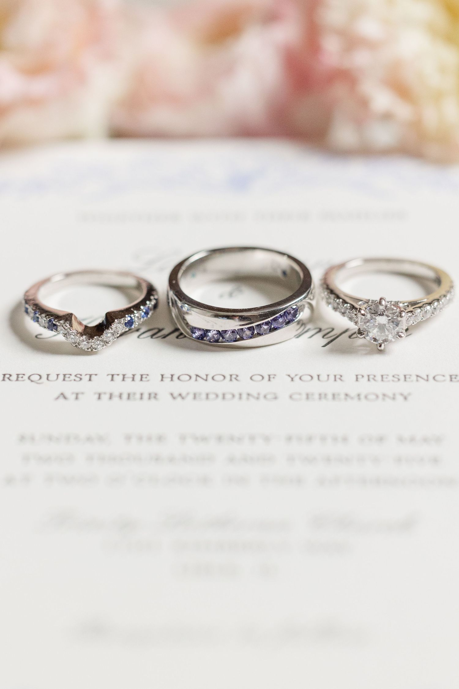 Three elegant diamond wedding rings laid on a white wedding invitation with romantic soft lighting.