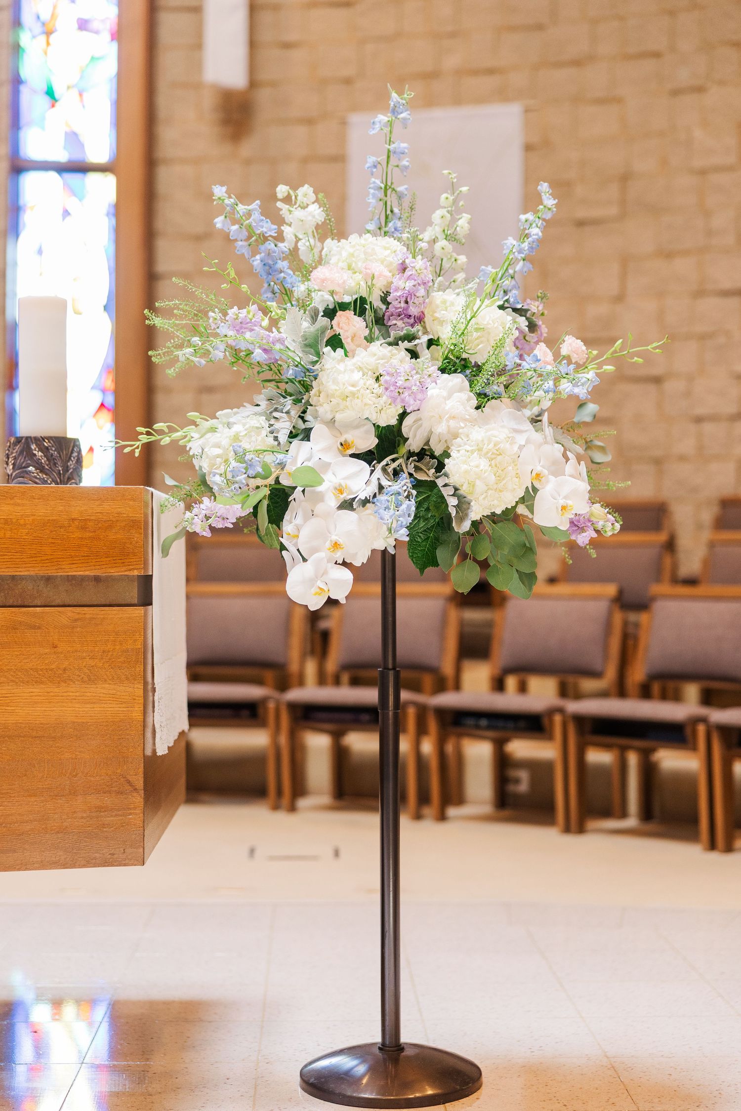 Elegant floral arrangements with white and purple flowers rest on wooden stands in a church sanctuary setting.