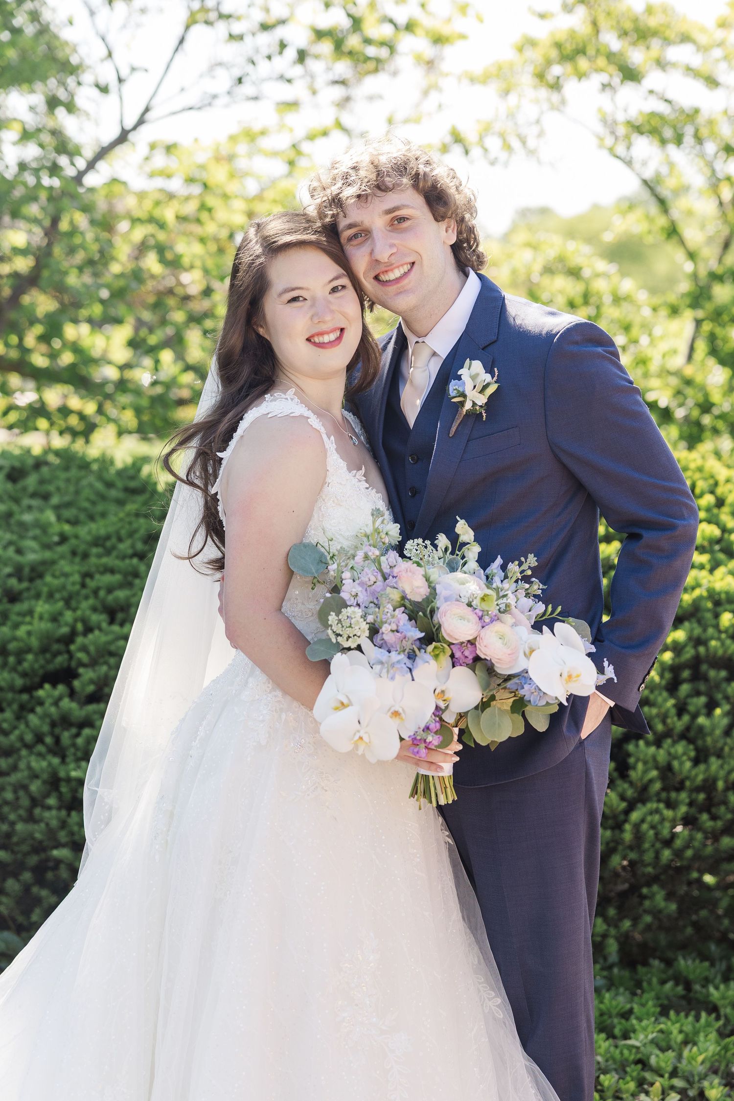Newlywed couple stands together outdoors with bride holding pastel pink and purple floral bouquet.