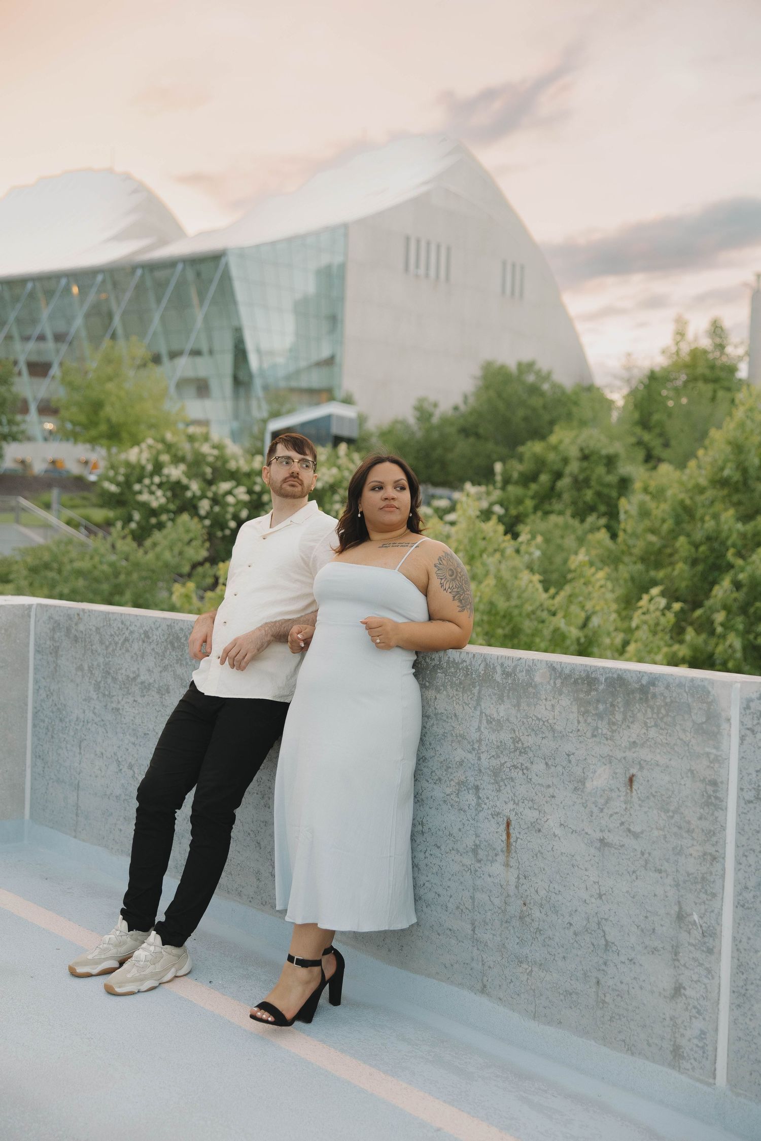 A couple poses on a concrete ledge with a modern glass building and greenery in the background at sunset.