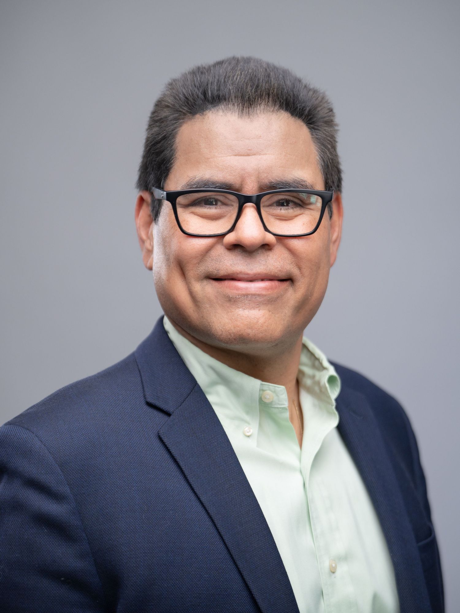 Professional headshot of a person wearing glasses, a light mint green dress shirt and navy blazer against a gray background.