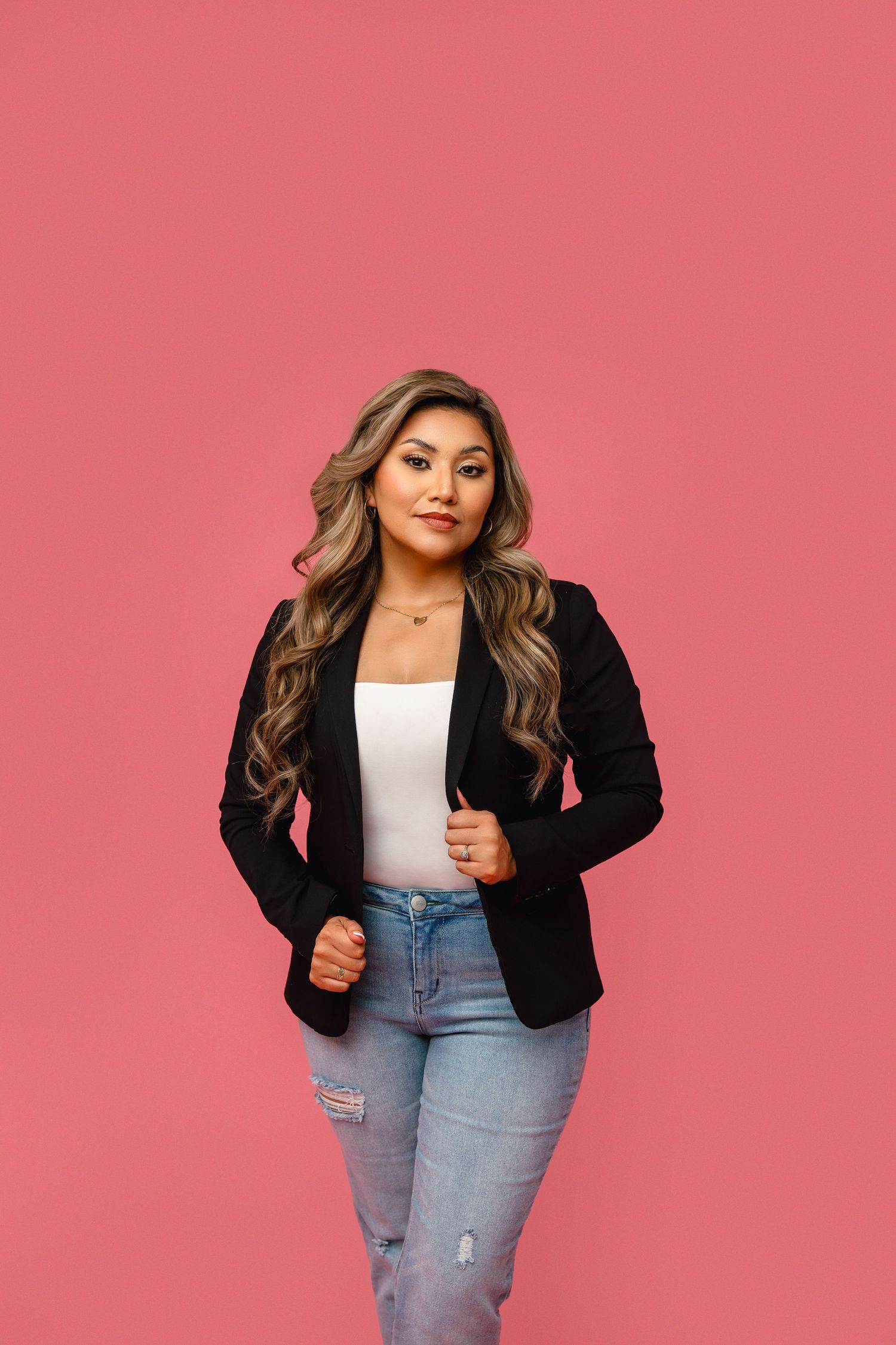 Professional portrait against pink background wearing black blazer, white top and distressed jeans.