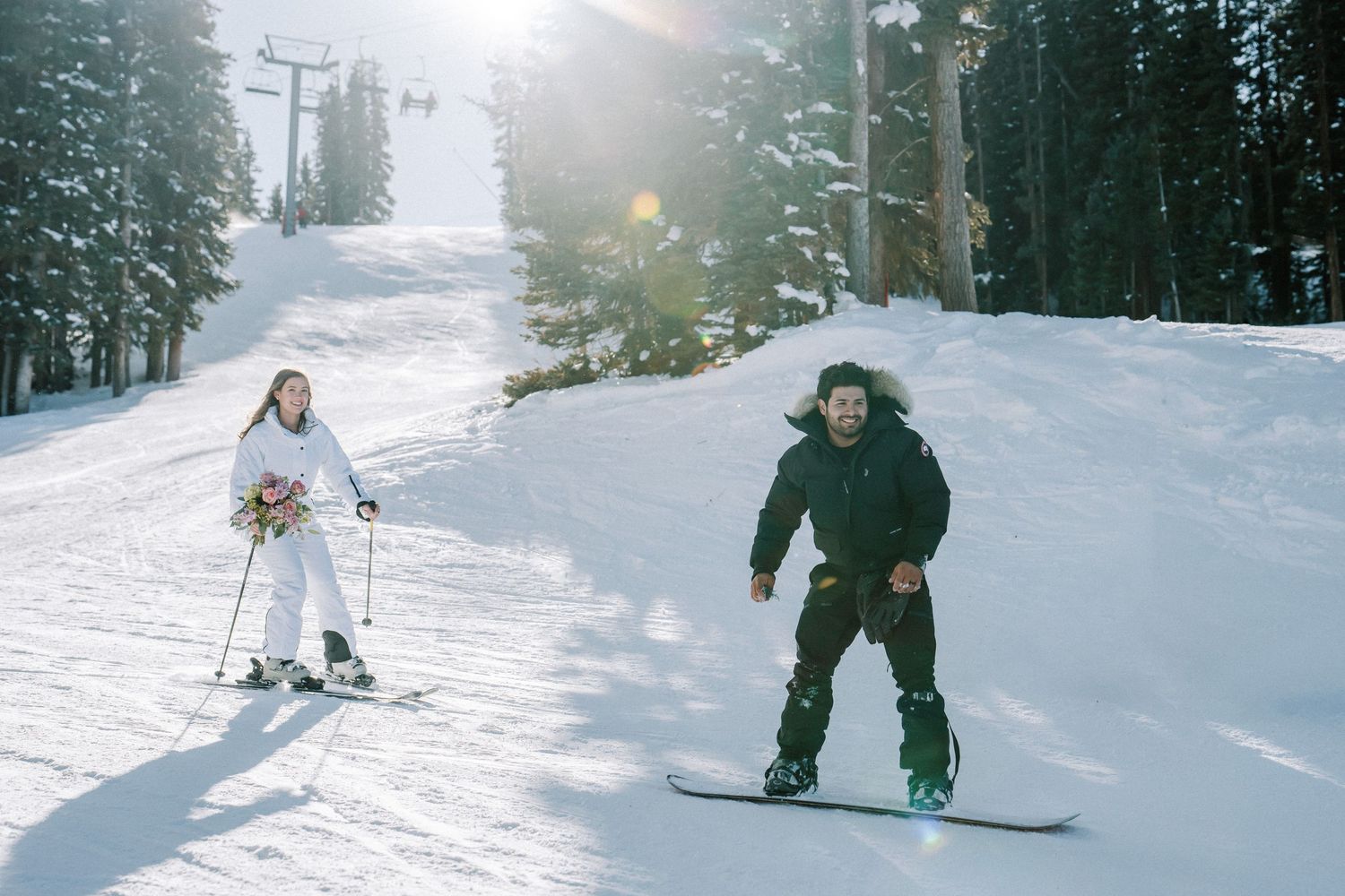 Two skiers ride down a snowy ski slope surrounded by evergreen trees on a sunny winter day.