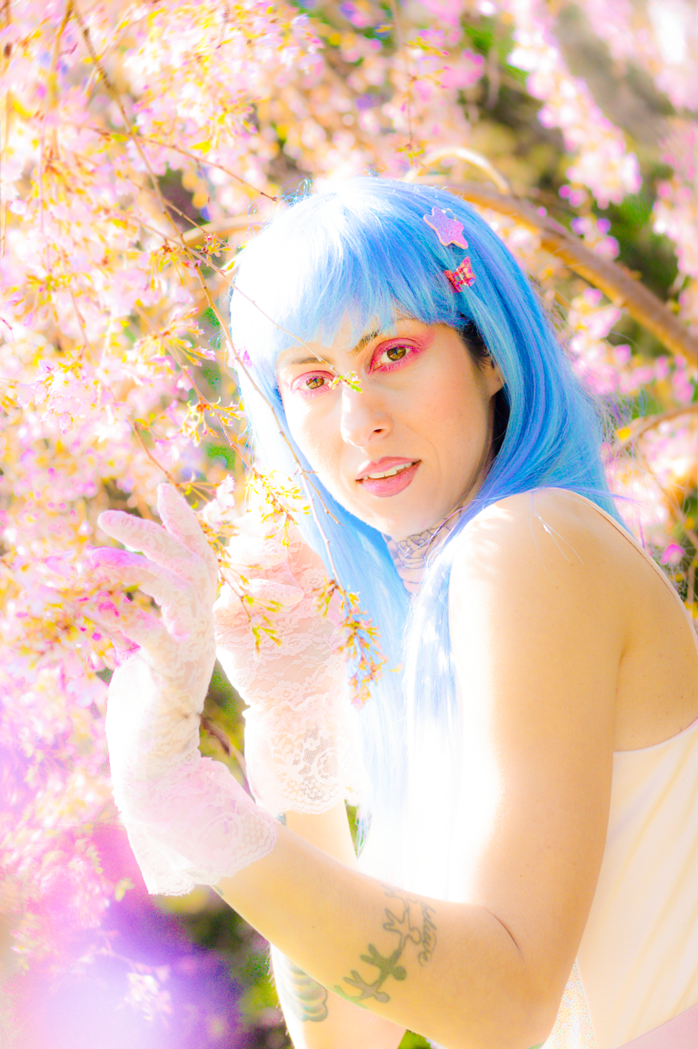 Dreamy portrait with blue hair and pink cherry blossoms creating an ethereal spring atmosphere in soft lighting.