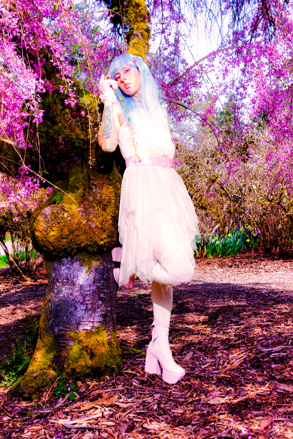 A person in a light pink dress and blue hair poses beneath blooming cherry blossom trees in an ethereal garden setting.