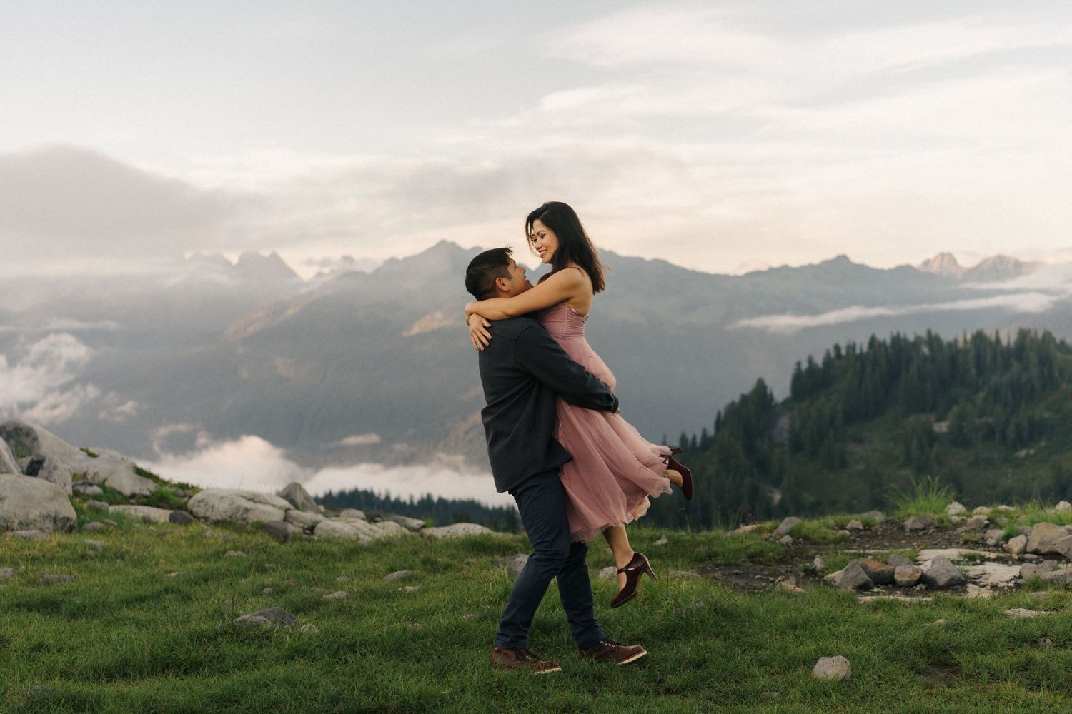 Two hands reaching to exchange something against a backdrop of majestic mountain peaks and evergreen forest at sunset.