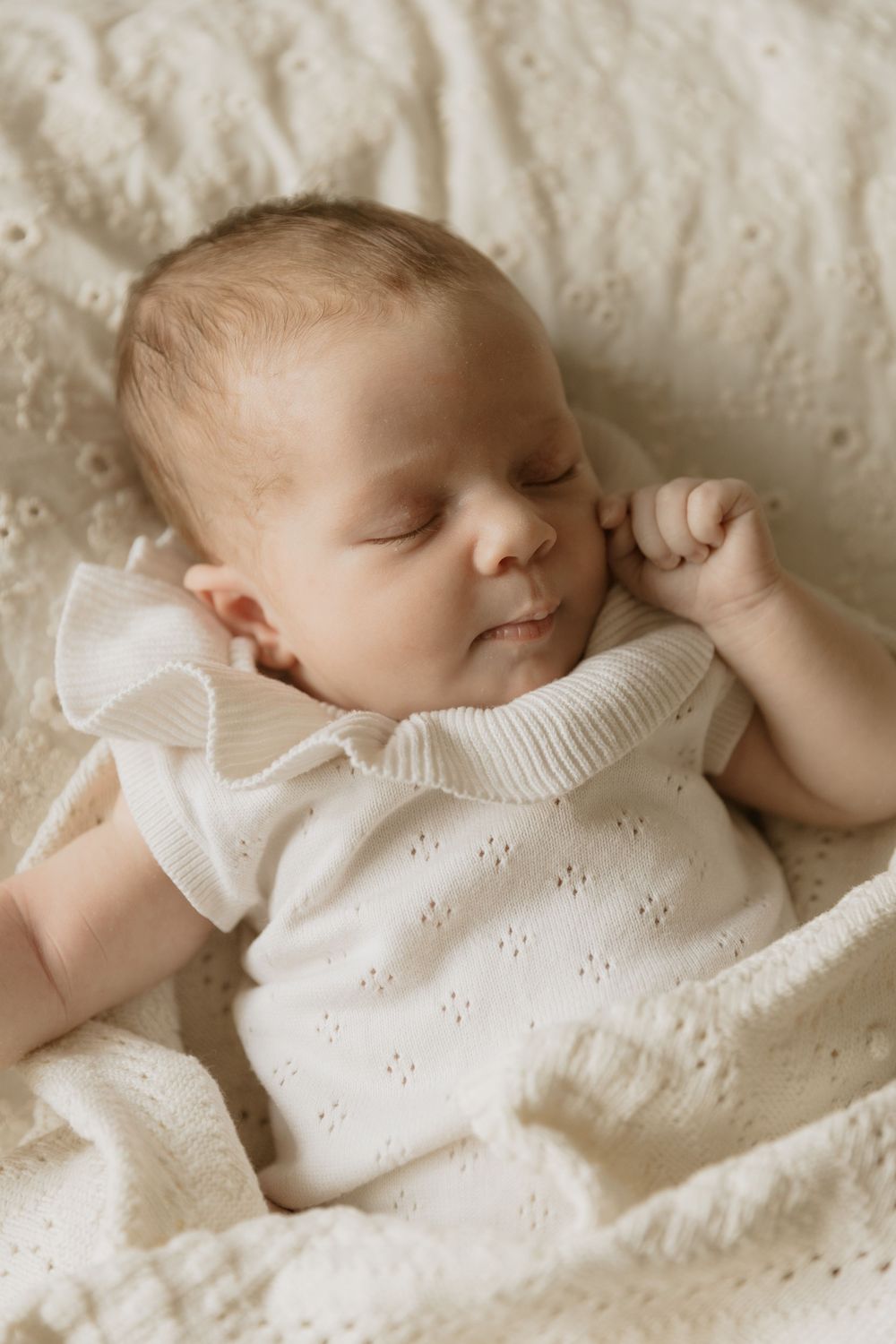 A sleeping baby wearing a white knit sweater lying on cream colored textured bedding.