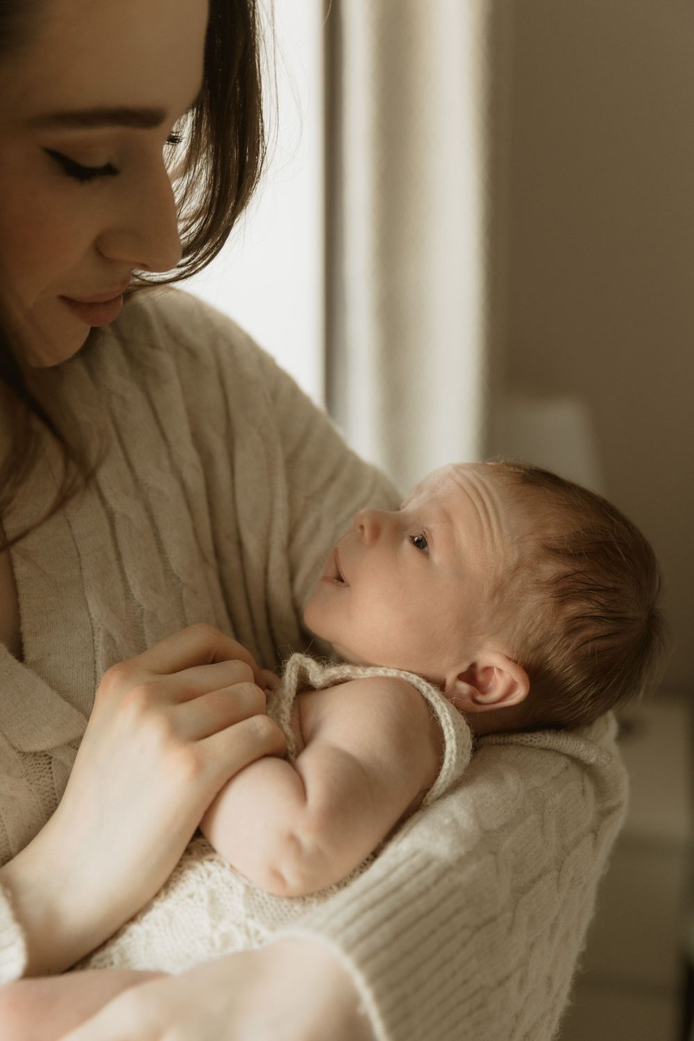 A mother in a beige sweater breastfeeding her baby in natural window light, shown in a series of tender poses.