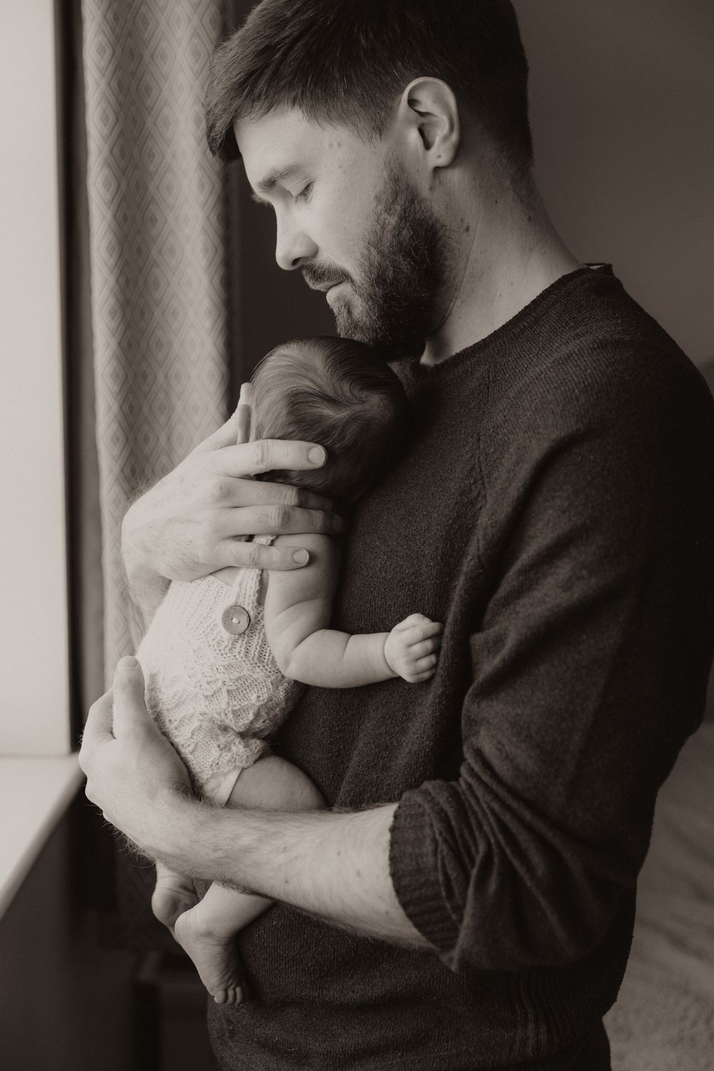 A tender moment captured in black and white as a parent in a sweater holds and gazes lovingly at their infant by a window.